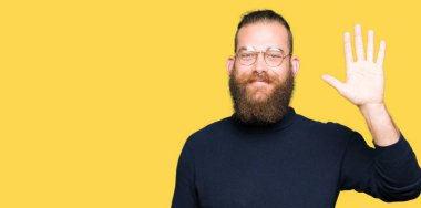 Genç Sarışın giyen gözlük ve balıkçı yaka kazak Waiving selam söyleyerek mutlu adam ve gülümseyen, dostça jest hoş geldiniz