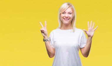 Gösterilen ve parmakları ile işaret izole arka plan üzerinde beyaz t-shirt giyen genç güzel sarışın kadın kendinden emin ve mutlu gülümseyerek süre yedi numara.