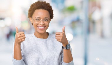 Young beautiful african american woman wearing glasses over isolated background success sign doing positive gesture with hand, thumbs up smiling and happy. Looking at the camera with cheerful expression, winner gesture. clipart