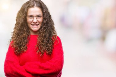 Güzel esmer kıvırcık saçlı Genç kız kameraya bakarak çapraz silah ile gülümseyen izole arka plan mutlu yüzüne gözlük ve kış kazak giyiyor. Olumlu bir kişi.