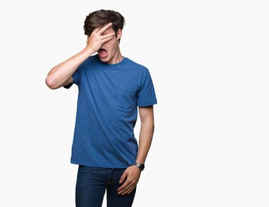Mahcup ifadeyle parmaklarımın arasından seyir yüz ve gözler el ile kapsayan şok bakmak izole arka plan üzerinde mavi t-shirt giyen genç yakışıklı adam.