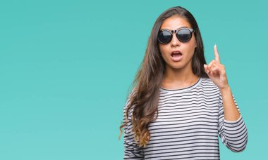 Genç güzel Arap kadın başarılı fikirle izole arka plan işaret parmağı üzerinde güneş gözlüğü takıyor. Çıktıktan ve mutlu. Bir numara.
