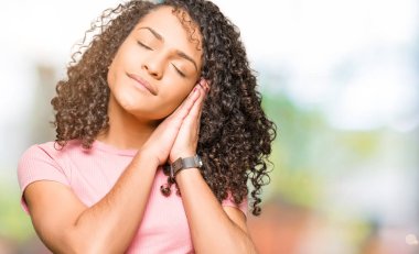 Rüya ve elleri ile kapalı gözler gülümseyen birlikte poz kıvırcık saçlı pembe uyku t-shirt giyen genç güzel kadın yorgun.