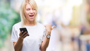 Genç güzel sarışın kadın işaret ve başparmak kadar mutlu yüz gülümseyen tarafı ile gösteren izole arka plan üzerinde akıllı telefon kullanma
