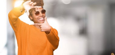 Güzel genç Afro-Amerikan kadın elle yapma çerçeve ve parmakları ile mutlu yüz gülümseyen izole arka plan üzerinde güneş gözlüğü takıyor. Yaratıcılık ve Fotoğrafçılık kavramı.