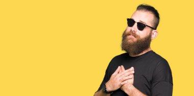 Genç hipster adam gözleri kapalı gözler ve yüz minnettar jest ile göğsünde eller ile gülümseyen güneş gözlüğü giyiyor. Sağlık konsepti.