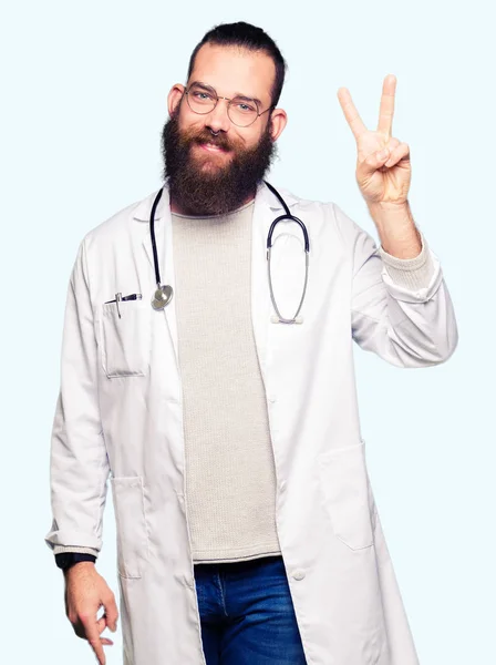 若いブロンド医師男性でひげを示す医療の着用のコートと自信を持って 幸せな笑みを浮かべている間 つ番号を指で上向き — ストック写真