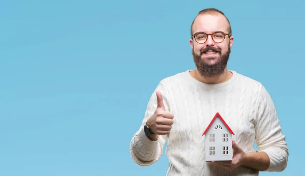 Young caucasian real state agent man holding house isolated background happy with big smile doing ok sign, thumb up with fingers, excellent sign