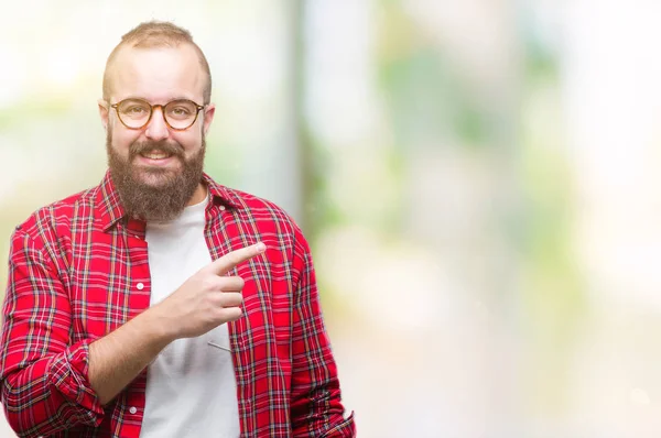 Young caucasian hipster man wearing glasses over isolated background cheerful with a smile of face pointing with hand and finger up to the side with happy and natural expression on face looking at the camera.