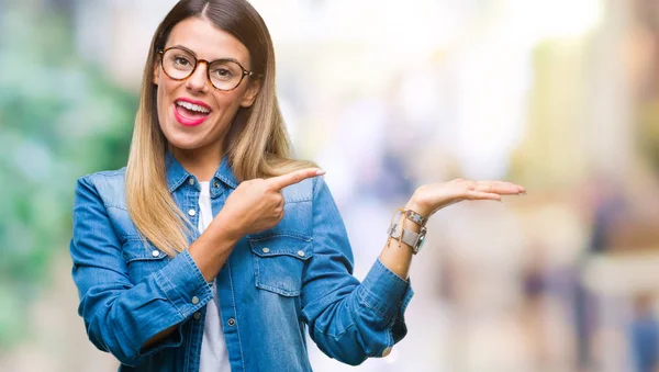 Young beautiful woman over wearing glasses over isolated background amazed and smiling to the camera while presenting with hand and pointing with finger.