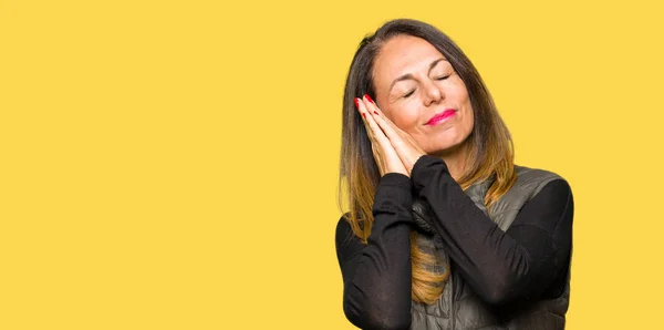 Beautiful middle age woman wearing winter vest sleeping tired dreaming and posing with hands together while smiling with closed eyes.