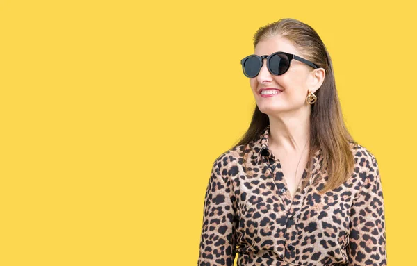 Middle age mature rich woman wearing sunglasses and leopard dress over isolated background looking away to side with smile on face, natural expression. Laughing confident.