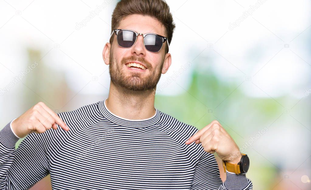 Young handsome man wearing sunglasses looking confident with smile on face, pointing oneself with fingers proud and happy.