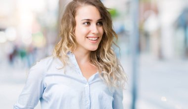 Beautiful young blonde business woman over isolated background looking away to side with smile on face, natural expression. Laughing confident.