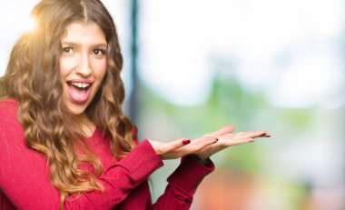 Kırmızı kazak giyen genç güzel kadın el ve açık avuç içi ile yan işaret, mutlu ve kendinden emin gülümseyen reklam sunan