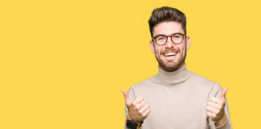 Gözlük başarı işareti el ile pozitif jest yapıyor giyen genç yakışıklı iş adamı başparmak gülümseyen ve mutlu. Neşeli bir ifadeyle, kazanan jest kameraya bakıyor.