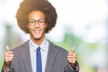El ile pozitif jest yapıyor gözlük başarı işareti giyiyor afro saçlı genç Afro-Amerikan iş adamı başparmak gülümseyen ve mutlu. Neşeli bir ifadeyle, kazanan jest kameraya bakıyor.