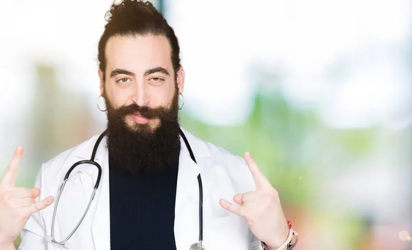 Doctor with long hair wearing medical coat and stethoscope shouting with crazy expression doing rock symbol with hands up. Music star. Heavy concept.