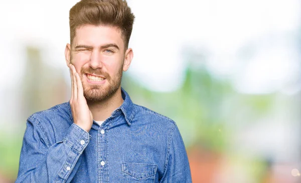 Young handsome blond man wearing casual denim shirt touching mouth with hand with painful expression because of toothache or dental illness on teeth. Dentist concept.