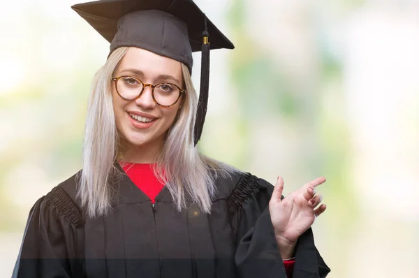 年轻的金发碧眼的女人穿着毕业制服 面带微笑 用手和手指指着一旁看着镜头 — 图库照片