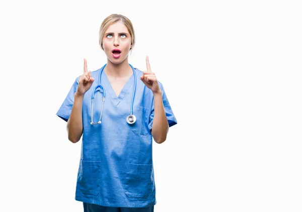 年轻美丽的金发医生外科医生护士妇女在孤立的背景惊讶和惊讶地抬起头来 用手指指指点点 举起双臂 — 图库照片