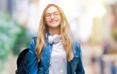 Yüzünde mutlu ve güzel bir gülümseme ile izole arka plan üzerinde kulaklık ve gözlük giyen genç güzel sarışın öğrenci kadın. Şanslı kişi.