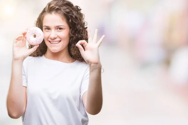 年轻的黑发女孩吃甜甜圈在孤立的背景做确定的标志与手指 优秀的象征 — 图库照片
