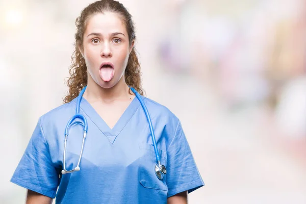 Young brunette doctor girl wearing nurse or surgeon uniform over isolated background sticking tongue out happy with funny expression. Emotion concept.