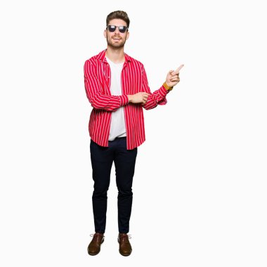 Kameraya bakarak yan el ve parmak ile işaret eden genç yakışıklı adam, yüzünde kocaman bir gülümseme ile güneş gözlüğü takıyor.