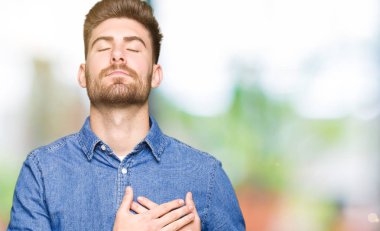 Casual denim ile gömlek gülümseyen giyen genç yakışıklı sarışın adam kapalı gözler ve yüzünde minnettar jest ile göğüs üzerinde eller. Sağlık kavramı.