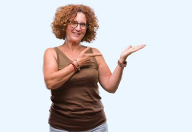 Orta yaş İspanyol kadın hayrete ve el ile sunulması ve parmak ile işaret ederken kameraya gülümseyen izole arka plan üzerinde gözlük.