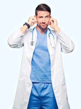 Yakışıklı Doktor adam yüksek sesle müzik gürültü rahatsız ifade parmaklarıyla kulaklarıyla kapsayan izole arka plan üzerinde tıbbi üniforma. Sağır kavramı.