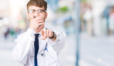 Genç doktor adam giyen hastane kat izole üzerinde arka plan sizin parmak el kamerasıyla ağzına, utanç ifadesi işaret eden gülme