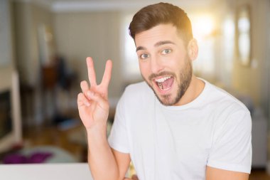 Mutlu yüz zafer işareti yaparken kameraya göz kırpıyor ile evde rahat beyaz t-shirt giyen genç yakışıklı adam gülümsüyor. İki numaralı.