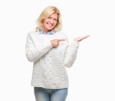 Hayret ve el ile sunulması ve parmak ile işaret ederken kameraya gülümseyen izole arka plan üzerinde kış kazak giyen orta yaş sarışın kadın.