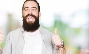 El ile olumlu hareketi yaparken uzun saç, sakal ve küpe başarı işareti ile genç adam başparmak gülümseyen ve mutlu. Neşeli bir ifadeyle, kazanan jest kameraya bakıyor.