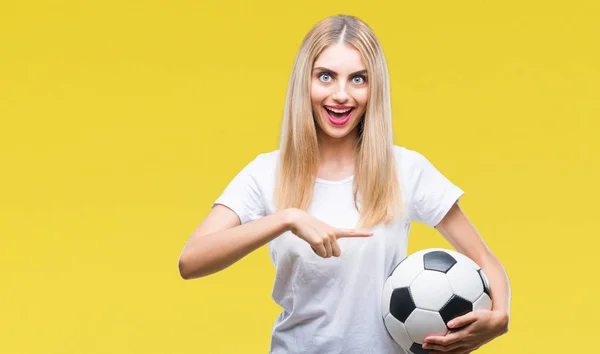 年轻美丽的金发女子持有足球在孤立的背景非常高兴地指着手和手指 — 图库照片