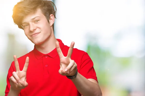勝利のサインをやって指を表示するカメラを笑みを浮かべて孤立の背景に赤い シャツを着ての若いハンサムな男 — ストック写真