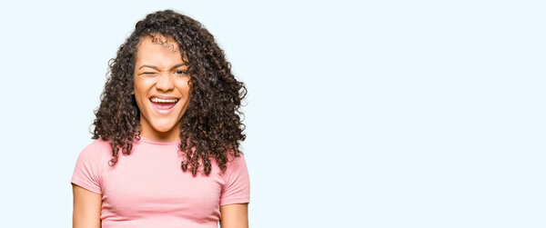 Young beautiful woman with curly hair wearing pink t-shirt winking looking at the camera with sexy expression, cheerful and happy face.