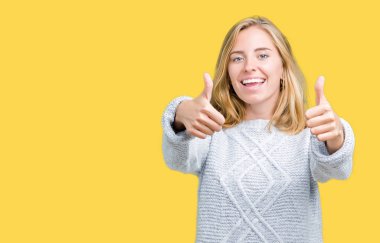 El ile pozitif jest yapıyor onaylama izole arka plan üzerinde kış kazak giyen güzel genç kadın başparmak gülümseyen ve başarı için mutlu. Kamera, kazanan jest arıyorsunuz.