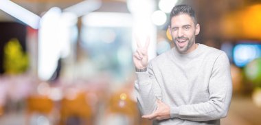 Mutlu yüz zafer işareti yaparken kameraya göz kırpıyor ile gülümseyen izole arka plan üzerinde sweatshirt giyen genç yakışıklı adam. İki numaralı.