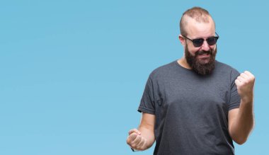 İzole arka plan kutlama üzerinde güneş gözlüğü takmış Genç beyaz hipster adam şaşırttı ve başarı için kaldırdı kollarını açarak hayret ve gözleri açık. Kazanan kavramı.