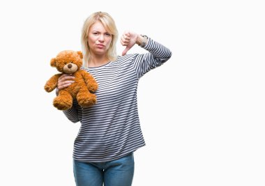 Kızgın yüz, eksi işareti aşağı antipati başparmak ile gösterilen, ret kavramı ile izole arka plan üzerinde oyuncak ayı peluş tutan genç güzel sarışın kadın