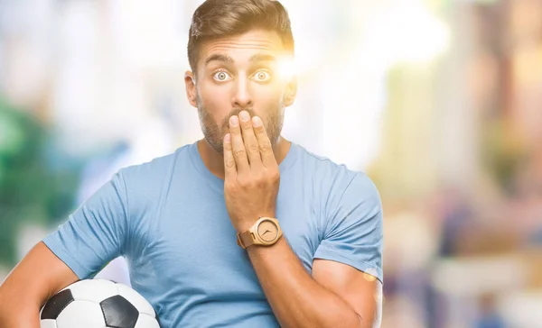 Young handsome man holding soccer football ball over isolated background cover mouth with hand shocked with shame for mistake, expression of fear, scared in silence, secret concept