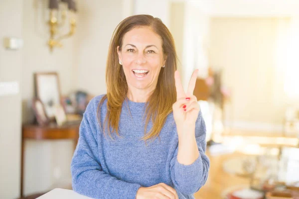 Beautiful middle age woman at home smiling with happy face winking at the camera doing victory sign. Number two.