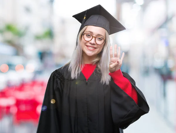 年轻的金发女子穿毕业制服在孤立的背景显示和指向与手指数字四同时微笑自信和快乐 — 图库照片