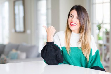 Genç güzel kadın giyiyor kış kazak evde seyir ve başparmak yanında işaret mutlu yüz ile gülümseyen.