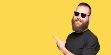 Yüzünde, kocaman bir gülümseme ile güneş gözlüğü takmış genç hipster adam kameraya bakarak yan el ve parmak ile işaret.