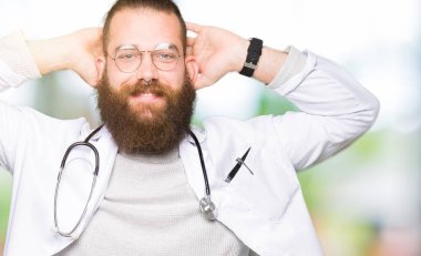 Tıbbi giyen sakallı genç sarışın doktor adam kat dinlenirken ve germe kollar ve eller baş ve boyun arkasında ile mutlu gülümseyerek