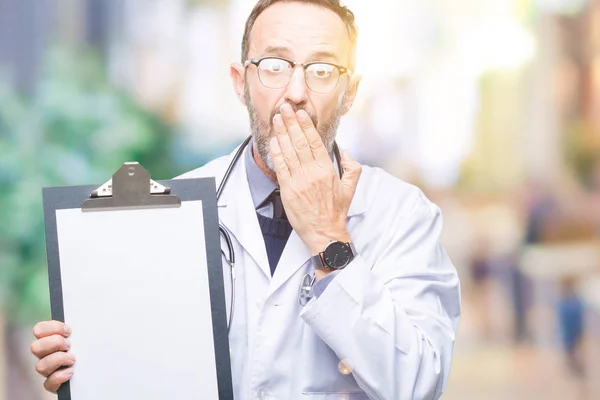 Middle age senior hoary doctor man holding clipboard isolated background cover mouth with hand shocked with shame for mistake, expression of fear, scared in silence, secret concept
